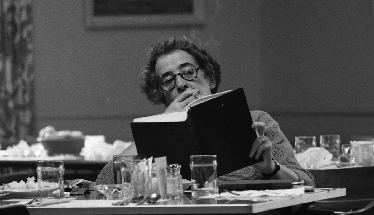 Hannah Arendt seated at a table, reading and thinking with a cigarette in her hand